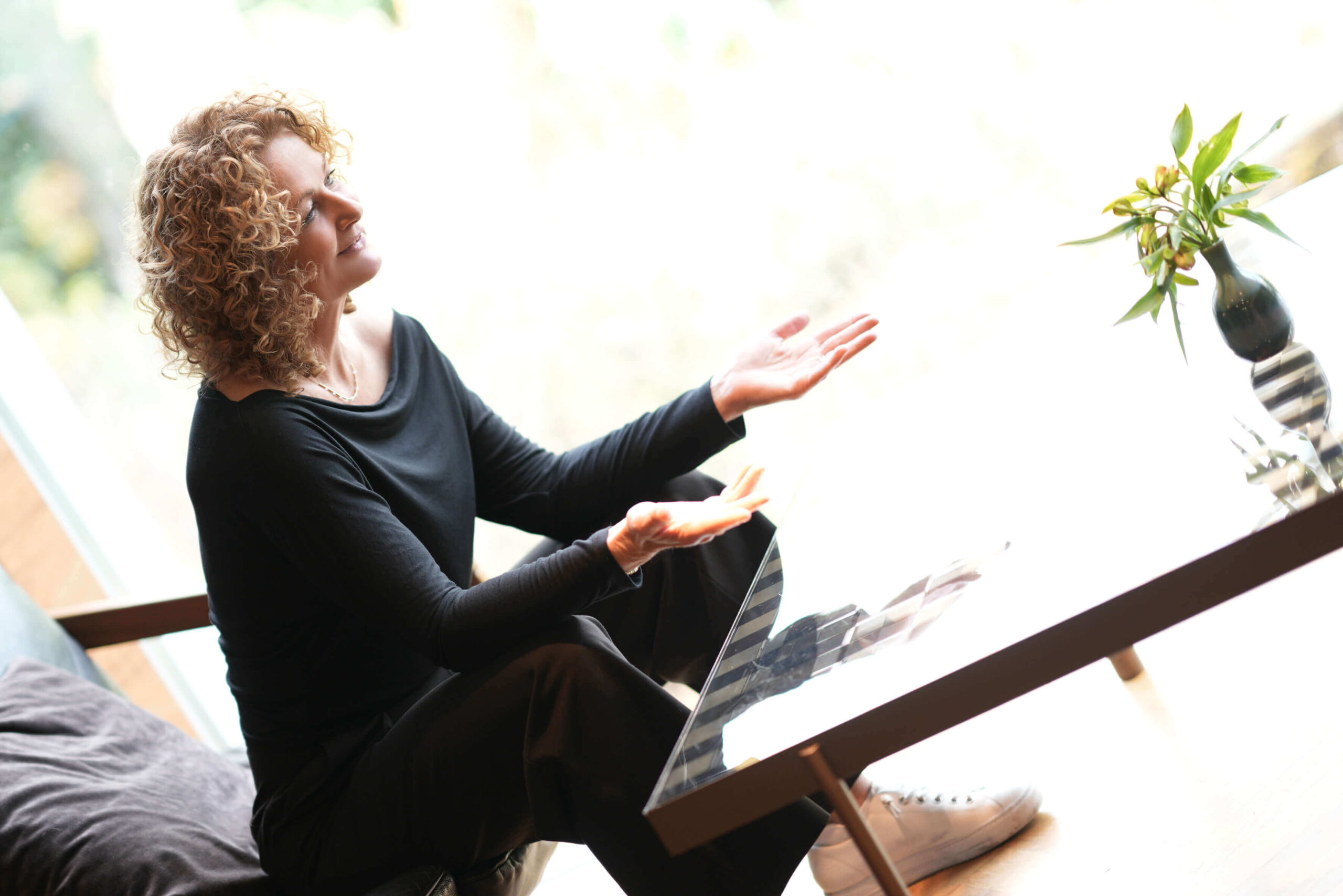 Alexandra Brindlinger sitzt an einem Tisch und spricht mit geöffneten Händen, als würde sie in einem ruhigen Gespräch Klarheit im Brandingprozess begleiten.